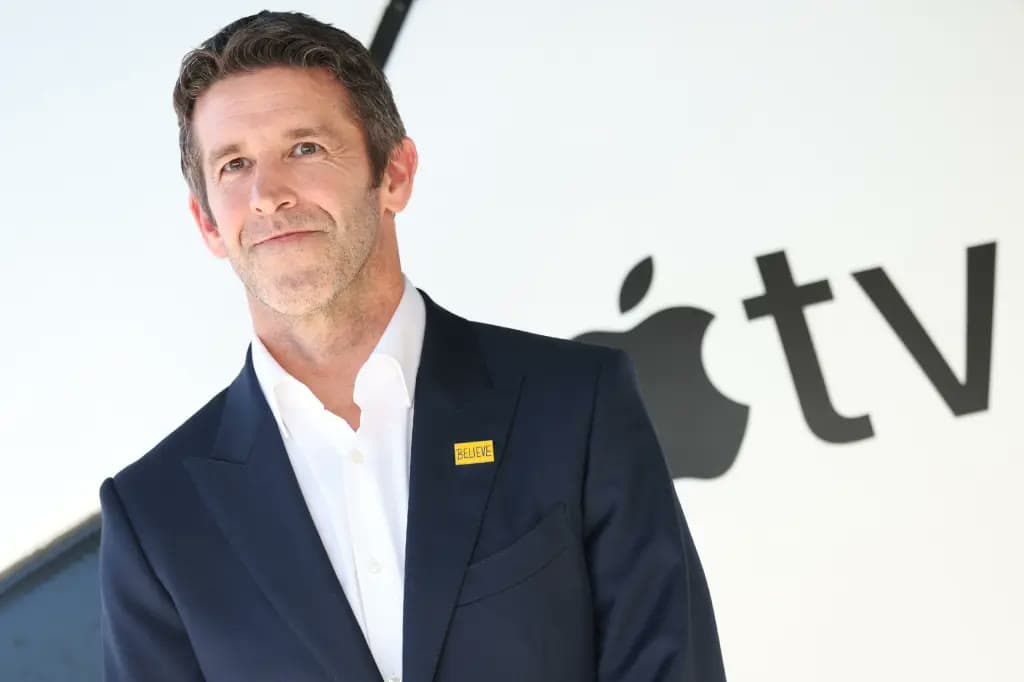 New Apple CEO John Ternus, wearing a navy suit with a yellow BELIEVE pin, stands in front of an Apple TV logo backdrop.