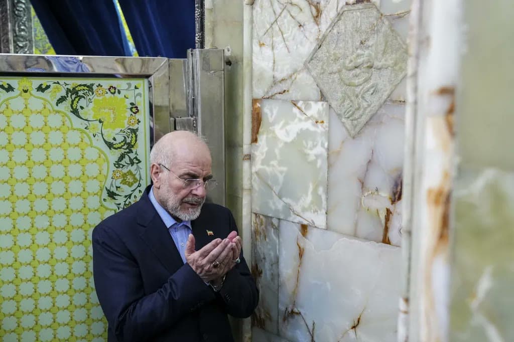 A bearded man in a suit prays with cupped hands at a marble shrine wall.