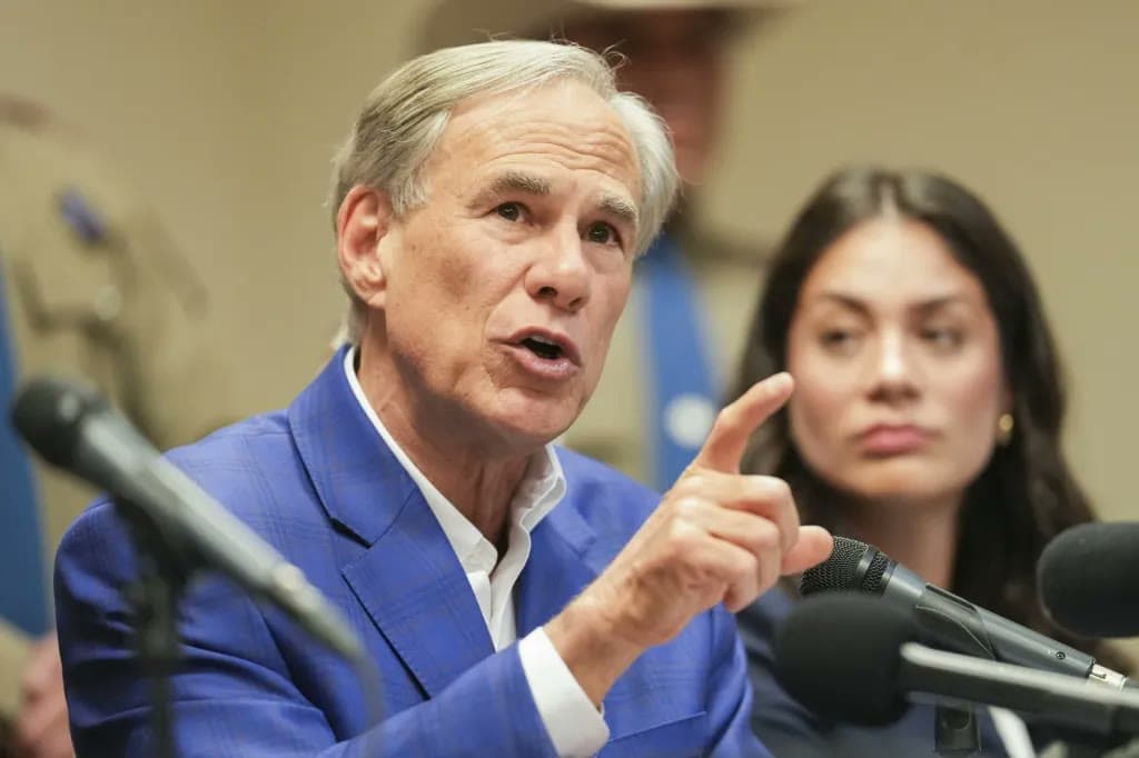Texas Governor Greg Abbott gestures while speaking at a podium with multiple microphones, with a woman looking on beside him.