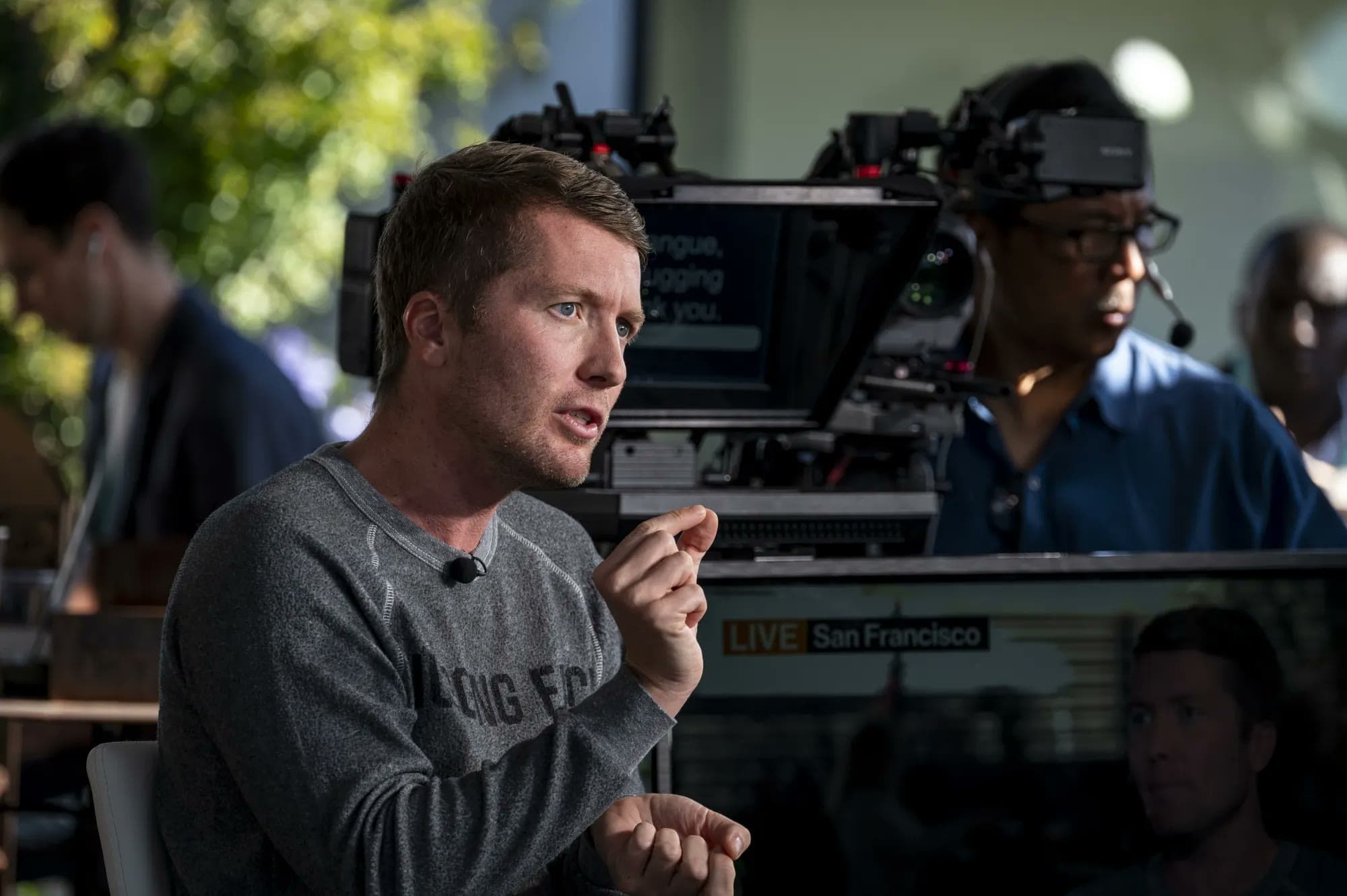 A man gestures while being interviewed on camera at an outdoor set with a 'LIVE San Francisco' broadcast monitor visible behind him.