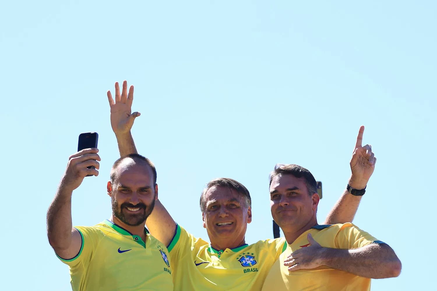 Three men in Brazil soccer jerseys wave and take a selfie against a clear blue sky.