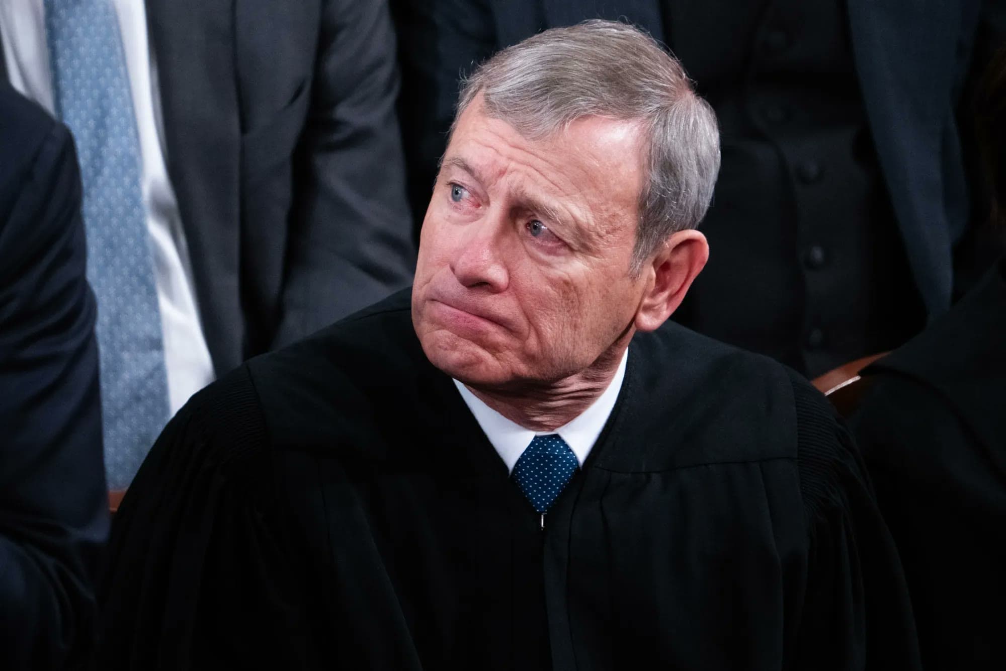 Chief Justice John Roberts, wearing judicial robes, looks to the side with a pensive expression at a formal event.