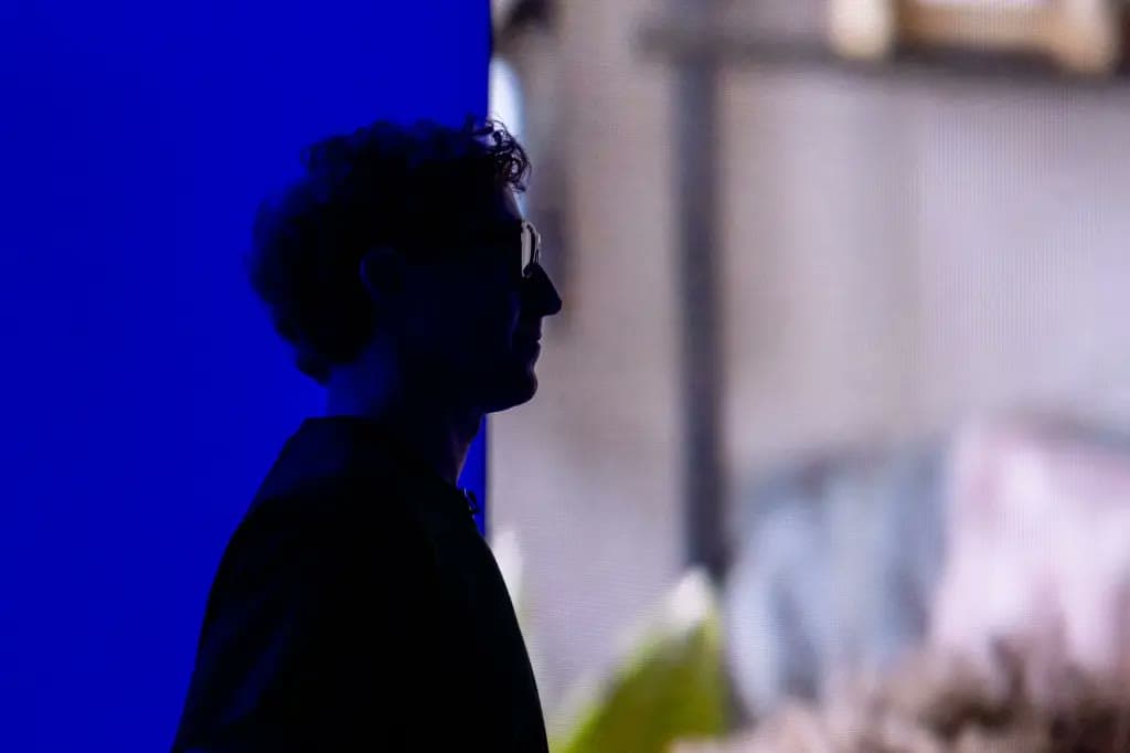 A silhouetted figure wearing glasses stands against a blue backdrop with a blurred building in the background.