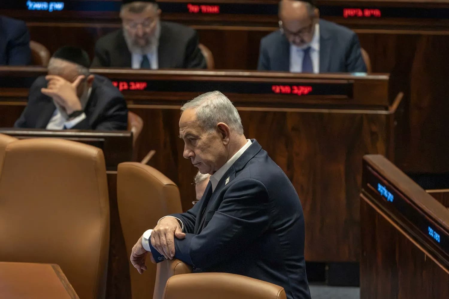 Israeli Prime Minister Benjamin Netanyahu sits in profile in the Knesset chamber, resting his arm on the seat back in front of him.