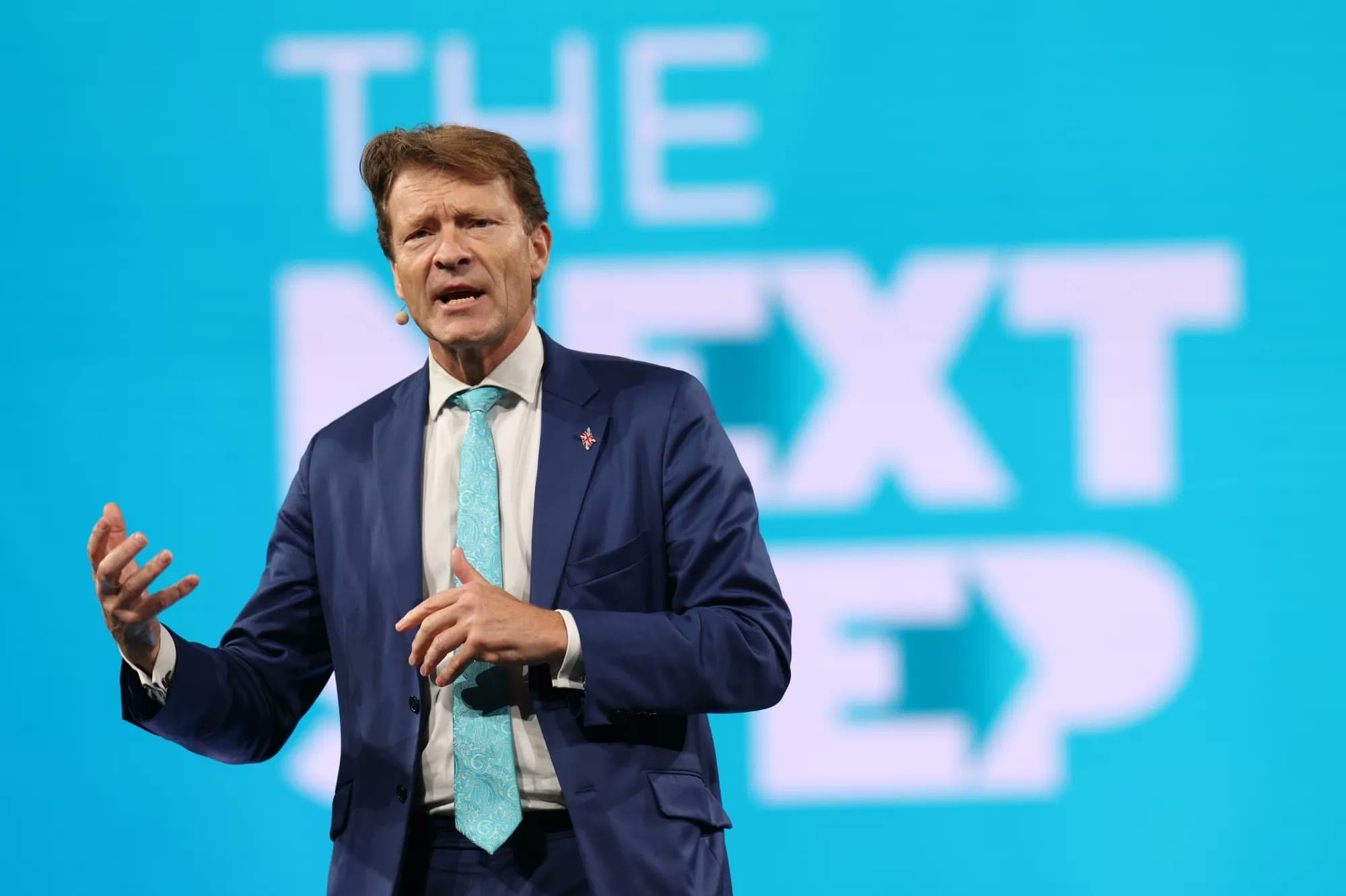 Richard Tice gestures while speaking on stage in front of a large teal screen reading 'THE NEXT STEP'.