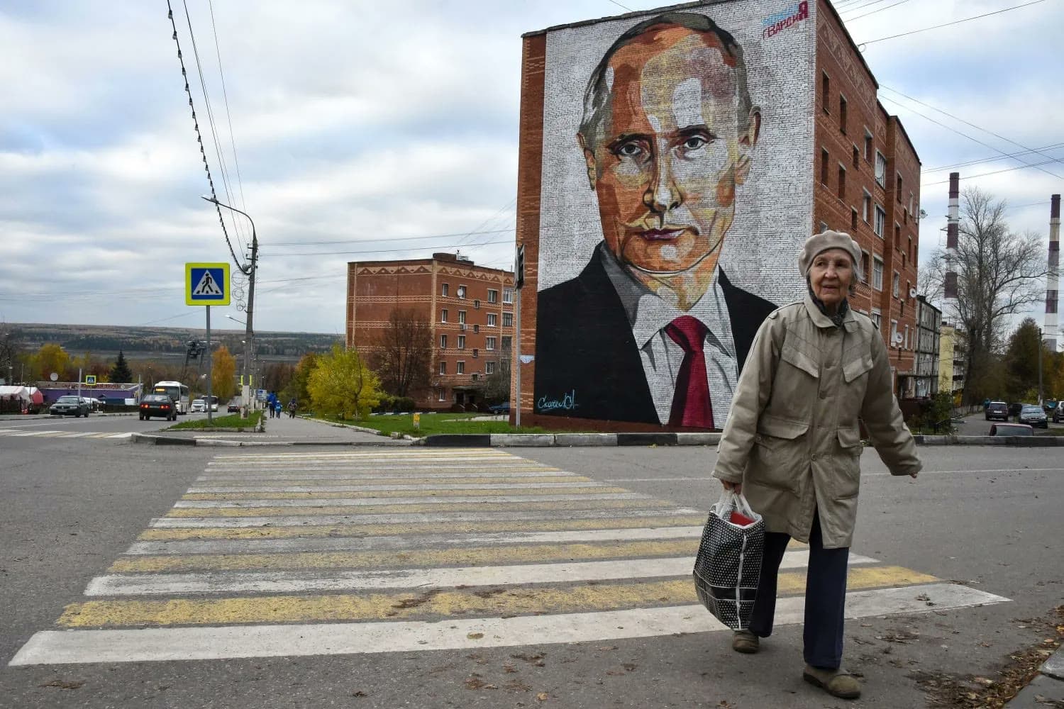 An elderly woman crosses a street in a Russian town beneath a large mural of Vladimir Putin painted on the side of an apartment building.