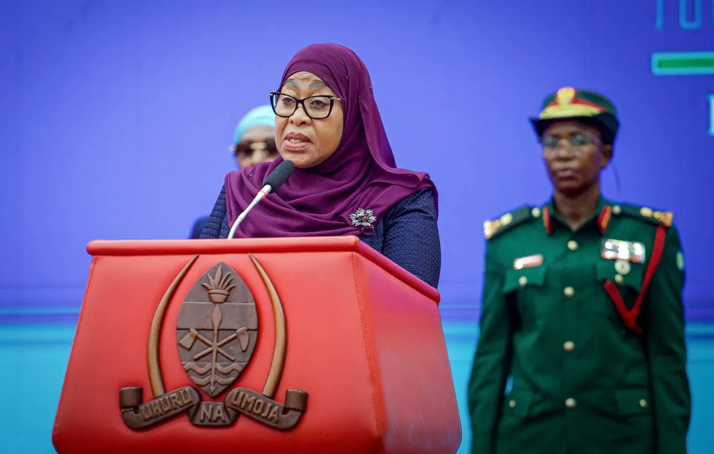 Tanzanian President Samia Suluhu Hassan speaks at a red podium bearing the national coat of arms, flanked by a uniformed military officer.