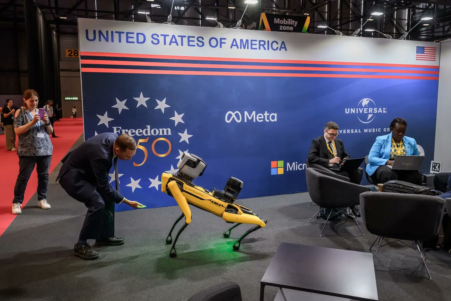 A man crouches beside a yellow robotic dog in front of a United States of America exhibit booth featuring Meta and Microsoft logos at a technology conference.