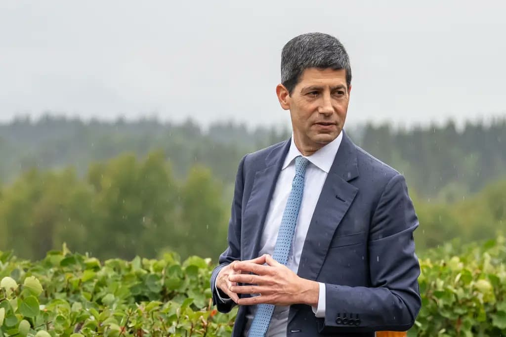 A man in a suit clasps his hands while speaking outdoors in front of green foliage on a rainy day.