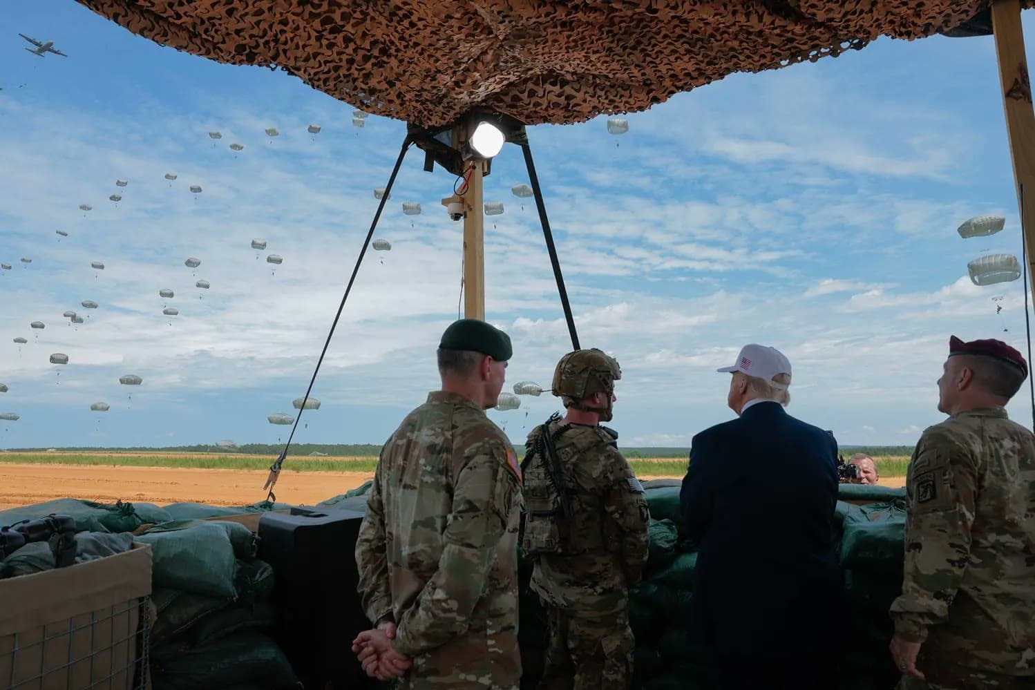 President Trump watches paratroopers descend under canopies during a military demonstration at Fort Bragg, flanked by soldiers under a camouflage canopy.