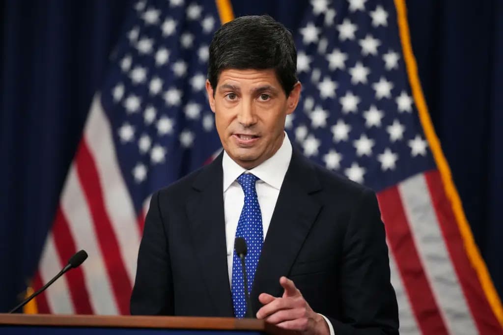 Kevin Warsh speaks at a podium in front of American flags with one hand raised mid-gesture.