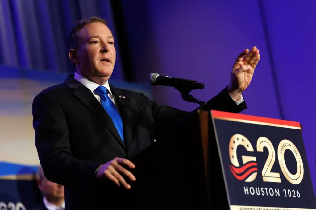 A man in a suit gestures while speaking at a podium bearing a G20 Houston 2026 sign.
