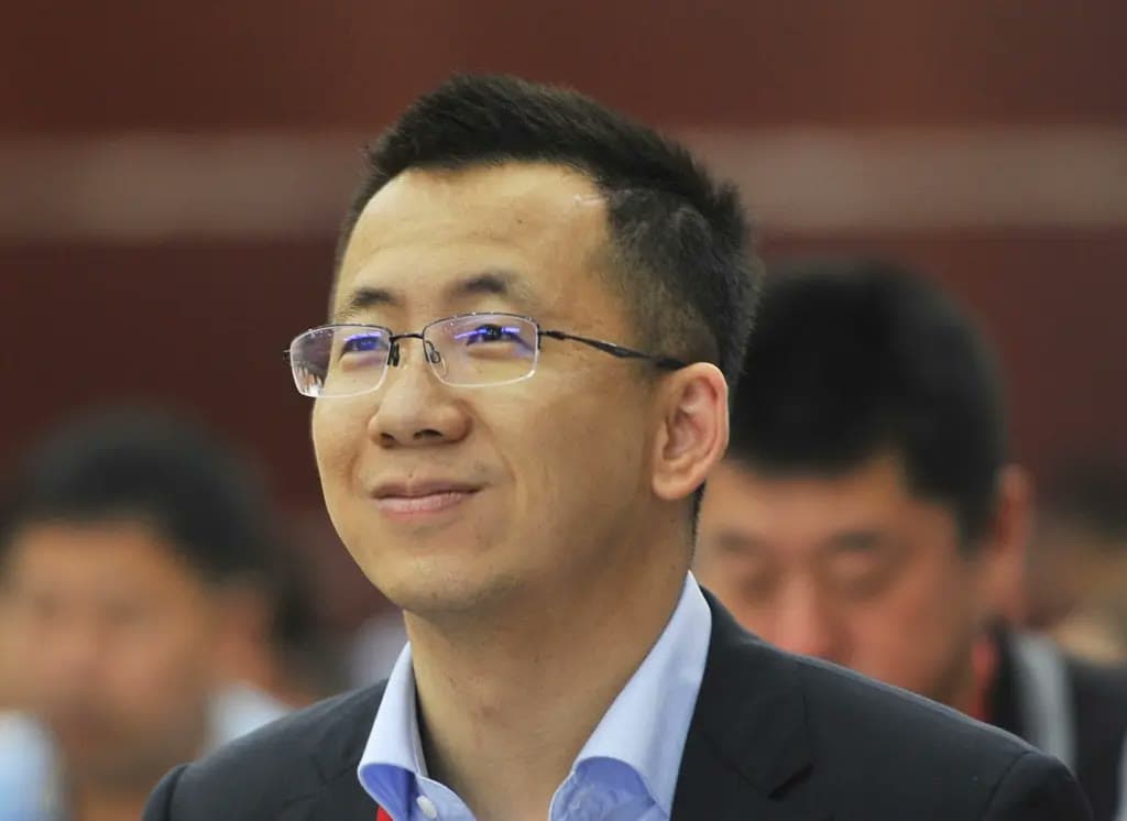 Zhang Yiming, wearing glasses and a dark suit, smiles while looking upward at an event.